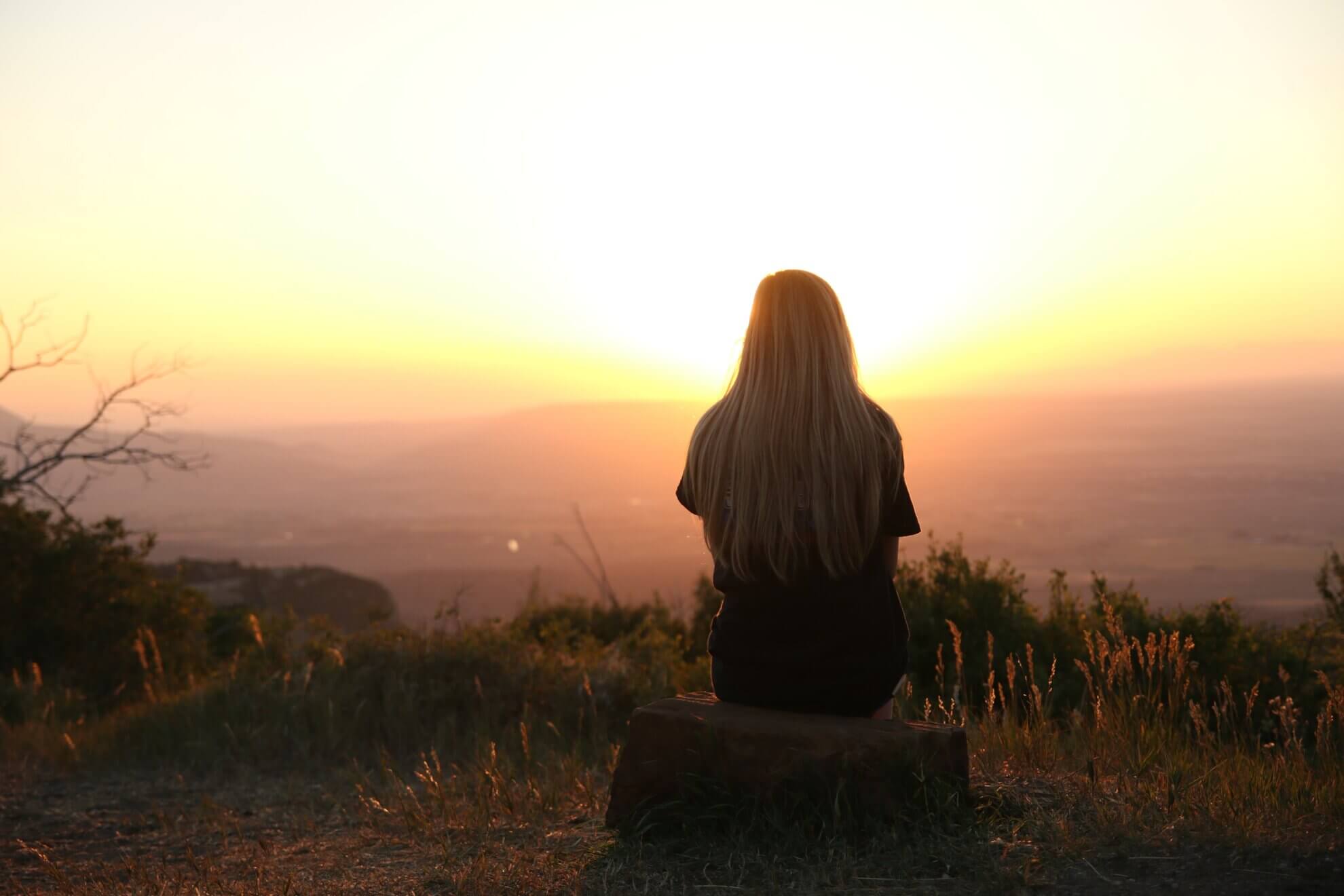 Eine Frau schaut auf einem Stein sitzend in den Sonnenaufgang. Diese Bild steht, dafür, dass es auch nach einer Dunklen Zeit weitergeht und man auch schwere Zeiten in einem individuellen Ritual verabschieden und hinter sich lassen kann.